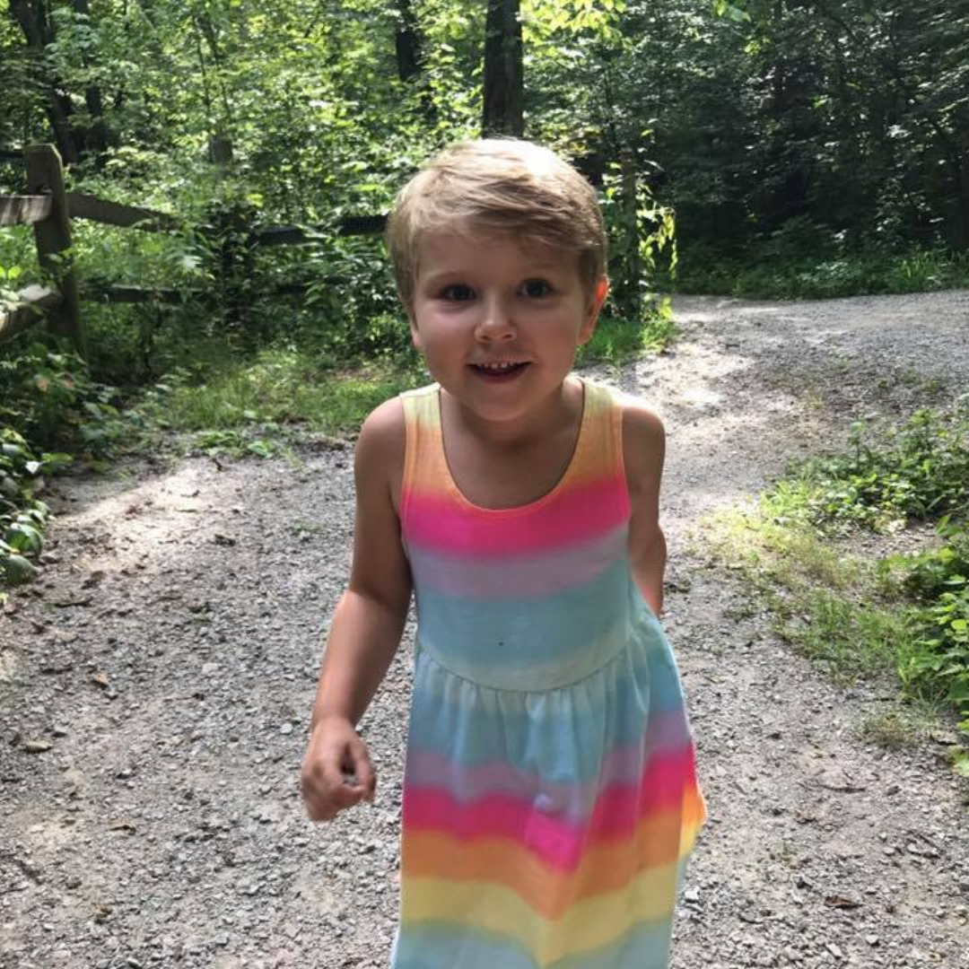A young child runs at the camera, smiling. They have short blonde hair and are wearing a rainbow dress. They're on a gravel trail, with a green summer forest behind them.